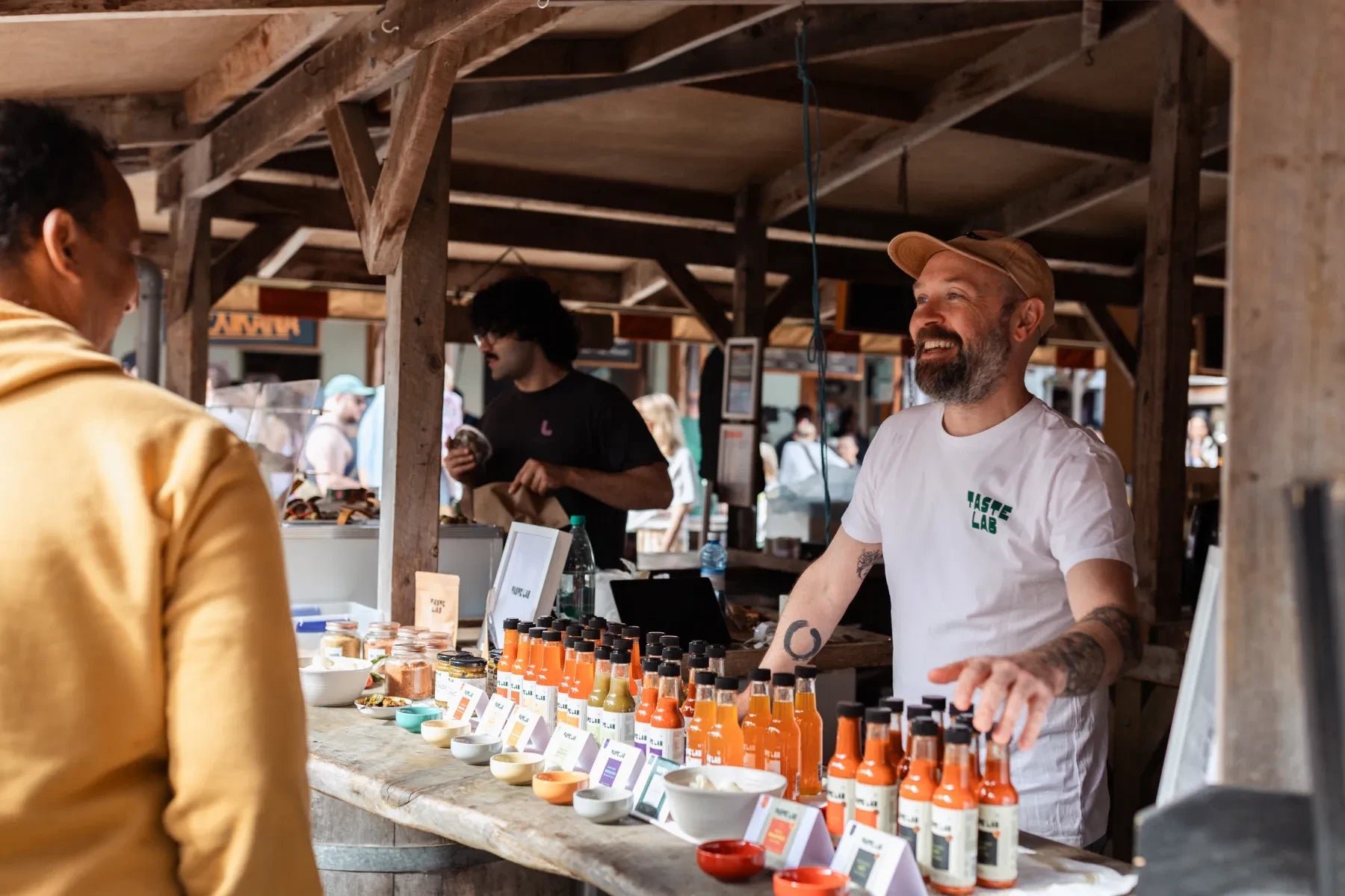 Taste Lab team member talking with a customer at a market stall, with rows of hot sauces and tasting bowls on the counter